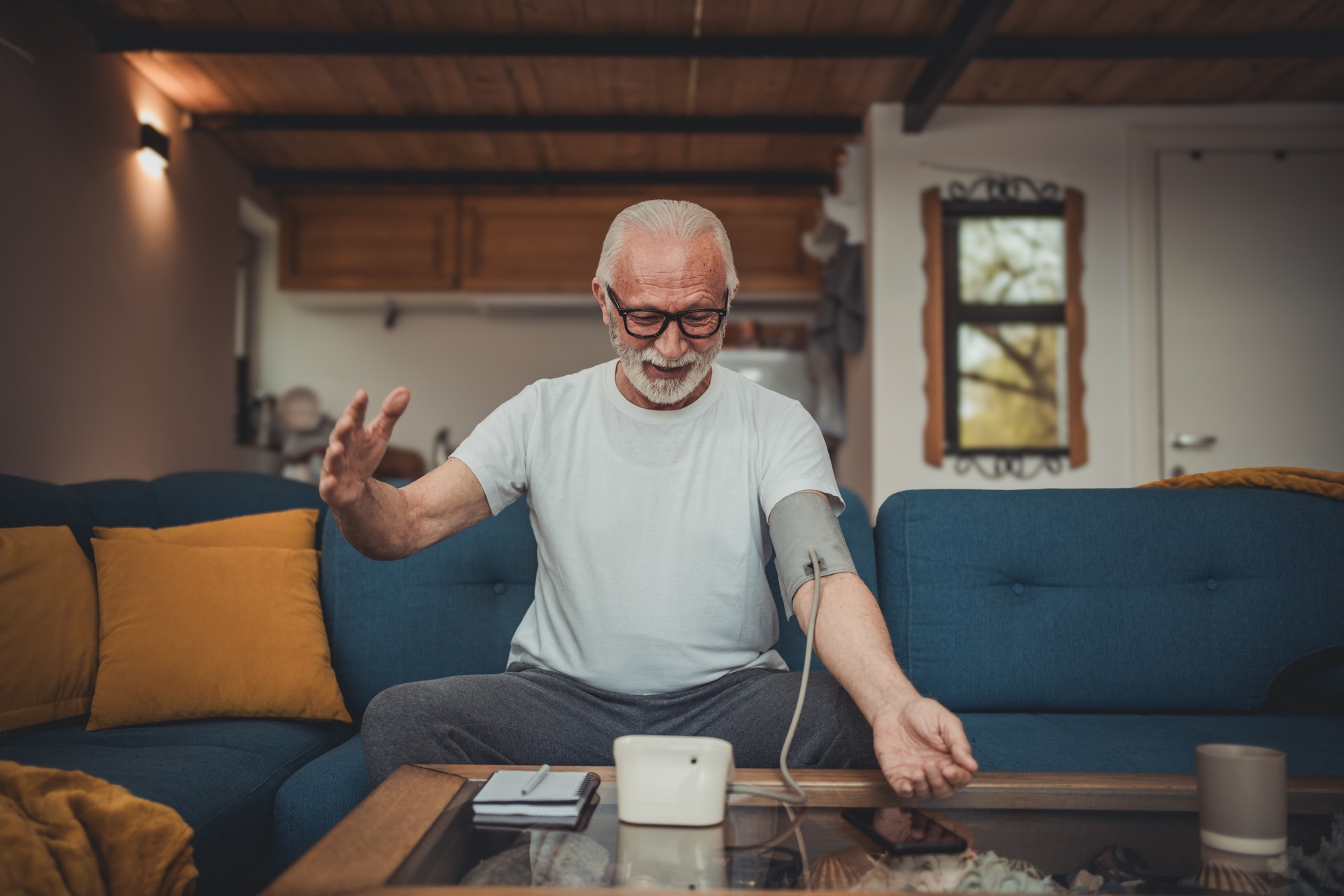 An elderly man measures his blood pressure with a digital meter while sitting on the sofa in his cottage